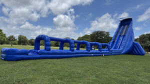 Blue inflatable obstacle course with a tall slide at the far end, set up on a grassy park field.